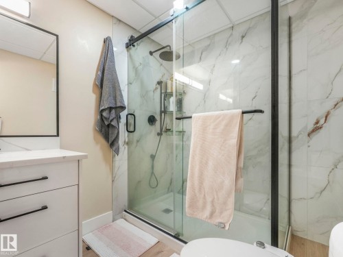 The bathroom features a modern vanity with a white countertop and black handles, a framed mirror, and a glass-enclosed shower with white marble-patterned tiles - 1147 Lincoln Crescent, Edmonton, AB - Indoor Photo Showing Bathroom
