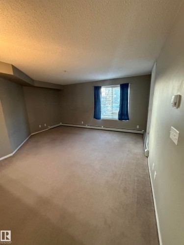 The property features a spacious room with carpeted flooring and a window providing natural light - 104 3207 James Mowatt Trail, Edmonton, AB - Indoor