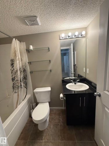 The bathroom features a bathtub with a shower, a toilet, and a vanity with a sink, black countertop, and dark cabinetry - 104 3207 James Mowatt Trail, Edmonton, AB - Indoor Photo Showing Bathroom