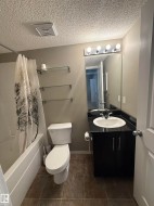 The bathroom features a bathtub with a shower, a toilet, and a vanity with a sink, black countertop, and dark cabinetry - 