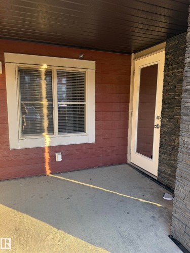 This property features a patio area with a concrete surface, bordered by red siding and a stone wall - 104 3207 James Mowatt Trail, Edmonton, AB - Outdoor With Exterior