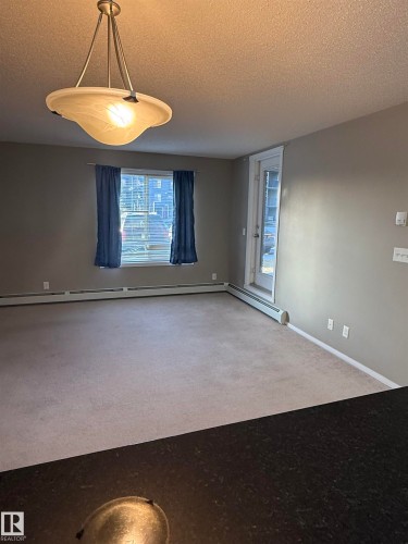 Living area featuring light-colored carpeting, a window with blinds and blue curtains, and a door leading outside - 104 3207 James Mowatt Trail, Edmonton, AB - Indoor Photo Showing Other Room