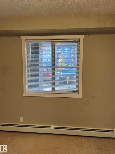 The property features a window with white blinds and trim, a wall-mounted electrical outlet, and baseboard heating - 104 3207 James Mowatt Trail, Edmonton, AB - Indoor Photo Showing Other Room