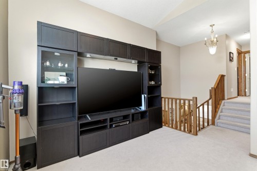 Living area featuring light-colored carpeting, a dark media console with display cabinets and shelving, and a wooden banister leading to a carpeted staircase - 442 Hunters Green, Edmonton, AB - Indoor Photo Showing Other Room