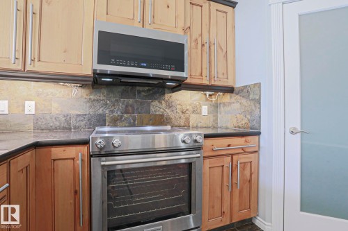 The kitchen features natural wood cabinetry, stainless steel appliances, a stone tile backsplash, and dark countertops - 205 79 Street Sw, Edmonton, AB - Indoor Photo Showing Kitchen