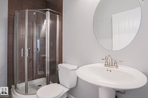 The bathroom features a corner glass shower enclosure with dark tiled walls, a white pedestal sink with a brushed nickel faucet, and a round wall mirror - 205 79 Street Sw, Edmonton, AB - Indoor Photo Showing Bathroom