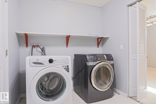 This utility area features a white front-loading washing machine and a dark gray front-loading dryer, alongside a white wall-mounted shelf with red brackets - 205 79 Street Sw, Edmonton, AB - Indoor Photo Showing Laundry Room