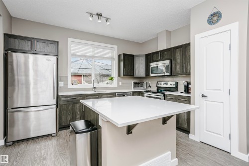 The kitchen features stainless steel appliances, dark wood-grain cabinetry, a white countertop island, and a window with blinds - 6116 Rosenthal Way, Edmonton, AB - Indoor Photo Showing Kitchen With Stainless Steel Kitchen With Upgraded Kitchen