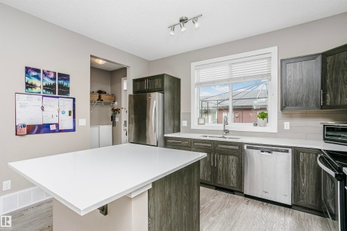 The kitchen features a white countertop island, dark wood grain cabinetry, a stainless steel dishwasher, and a window with blinds overlooking the exterior - 6116 Rosenthal Way, Edmonton, AB - Indoor Photo Showing Kitchen With Stainless Steel Kitchen