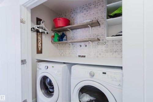 Dedicated laundry area featuring a countertop, two front-loading machines, and shelving with decorative patterned wall covering - 2878 Koshal Crescent, Edmonton, AB - Indoor Photo Showing Laundry Room
