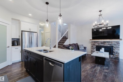 The open concept living area features dark wood flooring, a kitchen island with a double sink, stainless steel appliances, and a stone-clad fireplace - 2878 Koshal Crescent, Edmonton, AB - Indoor Photo Showing Kitchen With Double Sink With Upgraded Kitchen