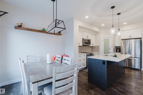 The open concept living area features a dining space with a contemporary linear chandelier, leading into a kitchen with a dark-toned island, light countertops, and stainless steel appliances - 2878 Koshal Crescent, Edmonton, AB - Indoor Photo Showing Kitchen With Upgraded Kitchen