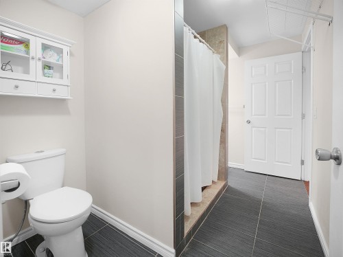 The bathroom features dark tiled flooring, a shower with a white curtain, and a white toilet - 8361 Shaske Crescent, Edmonton, AB - Indoor Photo Showing Bathroom