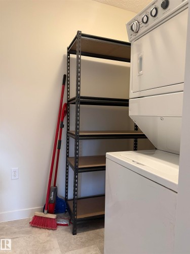 Dedicated laundry area featuring a stacked washer and dryer, a multi-shelf storage unit, and light-colored flooring - 331 10636 120 Street, Edmonton, AB - Indoor Photo Showing Laundry Room