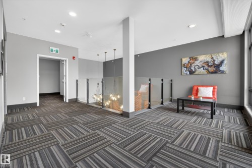This space features patterned carpet flooring, neutral colored walls, and a glass partition with industrial-style pendant lighting - 127 1154 Adamson Drive, Edmonton, AB - Indoor Photo Showing Other Room