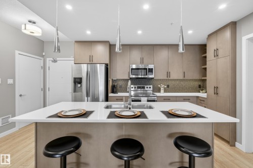 The kitchen features a spacious island with a white countertop and seating, stainless steel appliances, and extensive cabinetry for storage - 127 1154 Adamson Drive, Edmonton, AB - Indoor Photo Showing Kitchen With Stainless Steel Kitchen With Upgraded Kitchen