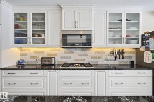 The kitchen features white cabinetry with glass-front upper cabinets, dark countertops, and a subway tile backsplash - 19 Escada Close, St. Albert, AB - Indoor Photo Showing Kitchen