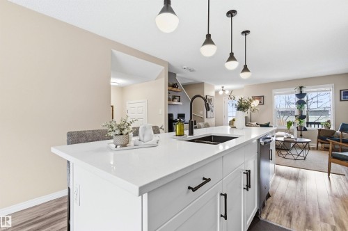 The kitchen features a white island with a sink and modern pendant lighting, overlooking a living area with hardwood style flooring and a large window - 3247 Cherry Crescent, Edmonton, AB - Indoor Photo Showing Kitchen With Double Sink