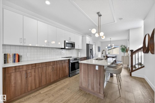 Contemporary kitchen featuring white upper cabinetry, wood grain lower cabinetry, stainless steel appliances, a white tile backsplash, and an island with a light-colored countertop - 7624 91 Avenue, Edmonton, AB - Indoor Photo Showing Kitchen With Upgraded Kitchen