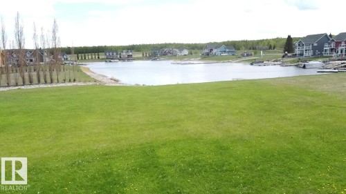 Expansive green lawn with a clear view of the lake and surrounding homes, featuring docks and a wooded area in the background - 13 Sunset Harbour, Rural Wetaskiwin County, AB 