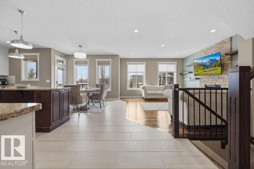 Open concept living area featuring light-toned flooring, a kitchen island with a granite countertop, a dining area with a chandelier, and a living room with hardwood flooring and a stone-accented wall - 76 Newgate Way, St. Albert, AB - Indoor