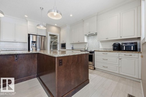 Spacious kitchen featuring an island with a light-colored countertop, white upper and lower cabinetry, stainless steel appliances, and light-colored tiled flooring - 76 Newgate Way, St. Albert, AB - Indoor Photo Showing Kitchen With Upgraded Kitchen