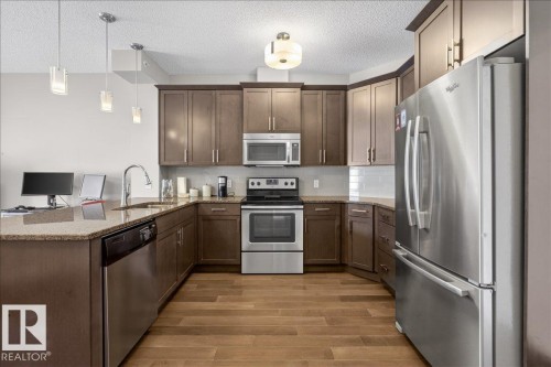 The kitchen features stainless steel appliances, wood cabinetry, granite countertops, and light wood flooring - 413 2203 44 Avenue, Edmonton, AB - Indoor Photo Showing Kitchen With Stainless Steel Kitchen