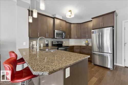 The kitchen features a spacious island with a granite countertop, dark wood cabinetry, stainless steel appliances, and wood flooring - 413 2203 44 Avenue, Edmonton, AB - Indoor Photo Showing Kitchen With Stainless Steel Kitchen With Double Sink