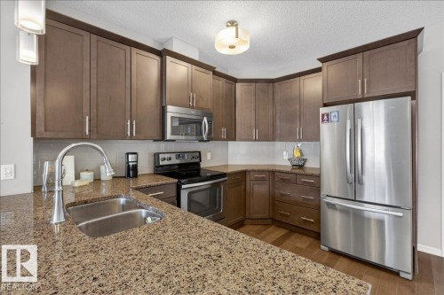 The kitchen features extensive cabinetry, granite countertops, a double basin sink with a gooseneck faucet, and stainless steel appliances including a refrigerator, microwave, and range - 413 2203 44 Avenue, Edmonton, AB - Indoor Photo Showing Kitchen With Stainless Steel Kitchen With Double Sink With Upgraded Kitchen