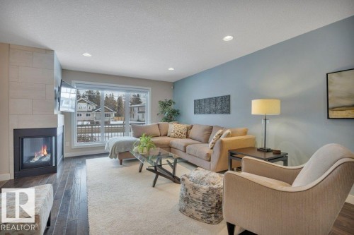 The living room features rich wood flooring, a gas fireplace with a light-toned tile surround, and a large window offering outdoor views - 9540 142 Street, Edmonton, AB - Indoor Photo Showing Living Room With Fireplace