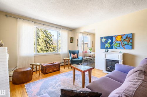 Living room featuring hardwood floors, a large window providing natural light, and a white fireplace - 12 6805 112 Street, Edmonton, AB - Indoor Photo Showing Living Room With Fireplace