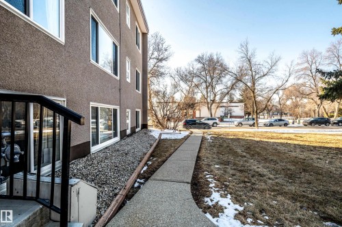 The property features a multi-story exterior with a textured finish and white-framed windows, complemented by a concrete walkway and surrounding trees - 12 6805 112 Street, Edmonton, AB - Outdoor With Exterior