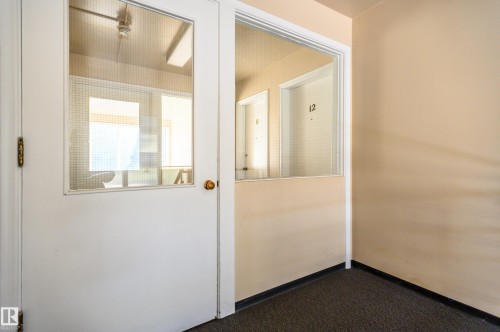 The property features a white door with a window and a brass door handle, complemented by beige walls and dark carpeting - 12 6805 112 Street, Edmonton, AB - Indoor Photo Showing Other Room