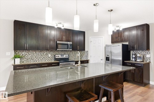 The kitchen features dark wood cabinetry, stainless steel appliances, a mosaic tile backsplash, and a large island with a gray countertop - 1668 Chapman Way, Edmonton, AB - Indoor Photo Showing Kitchen With Upgraded Kitchen