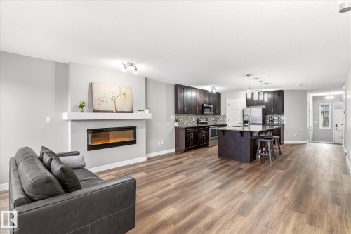 Expansive open-concept living area featuring hardwood flooring, a modern electric fireplace, and a kitchen with dark cabinetry, stainless steel appliances, and an island with bar seating - 1668 Chapman Way, Edmonton, AB - Indoor Photo Showing Living Room With Fireplace