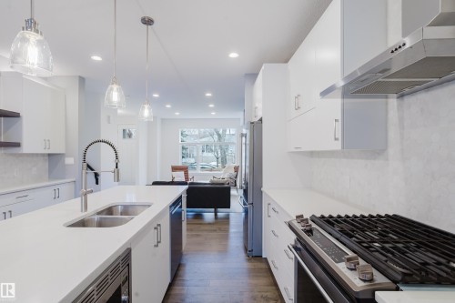 Well-appointed kitchen featuring white cabinetry, a large kitchen island with a double sink, stainless steel appliances, and sleek pendant lighting - 9612 72 Avenue, Edmonton, AB - Indoor Photo Showing Kitchen With Double Sink With Upgraded Kitchen