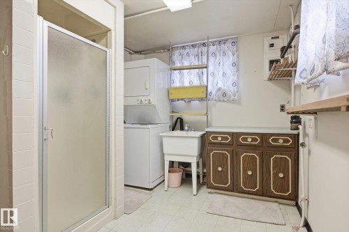 This practical space features a shower with a frosted glass door, a stacked washer and dryer, and a utility sink - 9131 74 Street, Edmonton, AB - Indoor Photo Showing Laundry Room