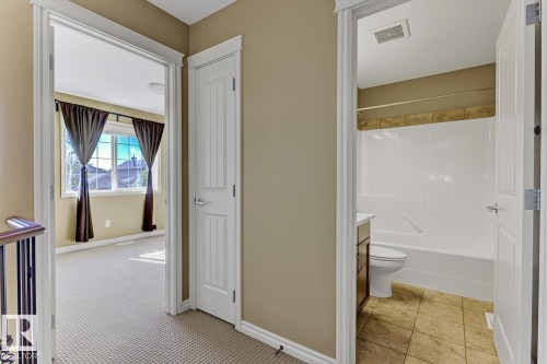 The property features a hallway with neutral carpeting and warm-toned walls, providing access to a room with large windows and a bathroom with tiled flooring and a tub/shower combination - 3105 Tredger Place, Edmonton, AB - Indoor Photo Showing Bathroom
