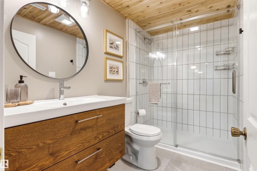 The bathroom features a wooden vanity with a white countertop and a round mirror, a glass-enclosed shower with white tiled walls, and a wood-paneled ceiling - 18315 68 Avenue, Edmonton, AB - Indoor Photo Showing Bathroom