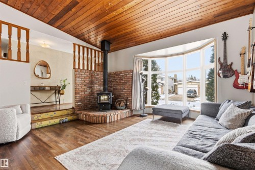 The living area features hardwood flooring, a wood-burning stove with a brick surround, and a bay window providing views of the neighborhood - 18315 68 Avenue, Edmonton, AB - Indoor Photo Showing Living Room With Fireplace