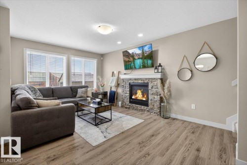 The living room features wood-look flooring, a stone-faced fireplace with a mantel, and two large windows with blinds - 16656 14 Avenue, Edmonton, AB - Indoor Photo Showing Living Room With Fireplace