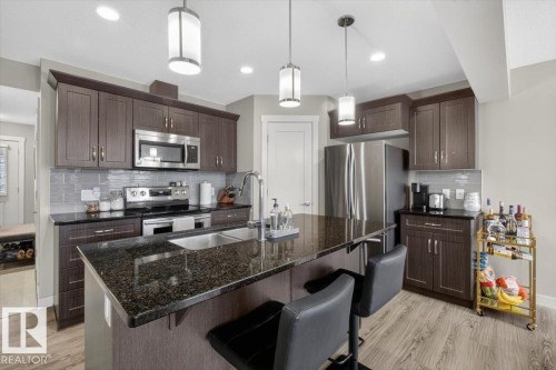The kitchen features dark wood cabinetry, stainless steel appliances, a dark granite island with an integrated sink, and pendant lighting - 16656 14 Avenue, Edmonton, AB - Indoor Photo Showing Kitchen With Stainless Steel Kitchen With Double Sink With Upgraded Kitchen