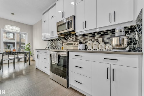 The kitchen features white cabinetry with black hardware, stainless steel appliances, a grey countertop, and a black and grey tiled backsplash - 32 1703 16 Avenue, Edmonton, AB - Indoor Photo Showing Kitchen With Upgraded Kitchen