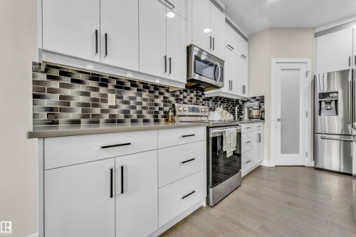 The kitchen features white cabinetry with dark hardware, a mosaic tile backsplash, and stainless steel appliances including a microwave, oven, and refrigerator - 32 1703 16 Avenue, Edmonton, AB - Indoor Photo Showing Kitchen With Upgraded Kitchen