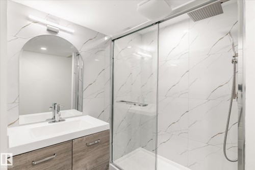 Contemporary bathroom featuring a vanity with a white countertop and wood-look cabinetry, an arched mirror, and a walk-in shower with clear glass enclosure and white tiled walls - 6540 184 Street, Edmonton, AB - Indoor Photo Showing Bathroom