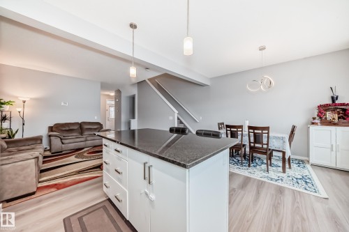 This open-concept main living area features light-colored flooring, a kitchen island with a dark countertop and white cabinetry, and a dining area with a modern light fixture - 5868 Anthony Crescent, Edmonton, AB - Indoor