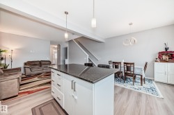 This open-concept main living area features light-colored flooring, a kitchen island with a dark countertop and white cabinetry, and a dining area with a modern light fixture - 