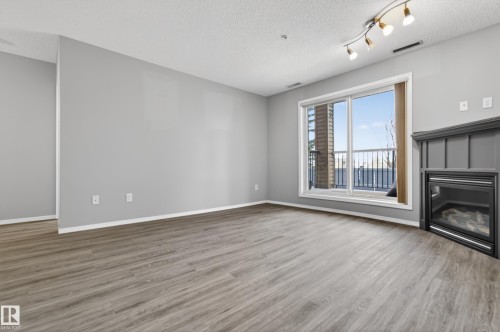 Living area featuring wood-look flooring, light grey walls, and a fireplace with a dark mantel - 131 2098 Blackmud Creek Drive Sw, Edmonton, AB - Indoor With Fireplace