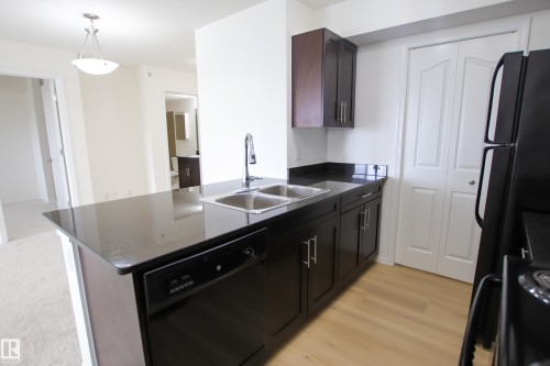 The kitchen features dark wood cabinetry, a double basin stainless steel sink, black countertops, and light wood flooring - 405 534 Watt Boulevard, Edmonton, AB - Indoor Photo Showing Kitchen With Double Sink