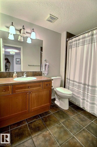 Bathroom featuring a wood vanity with a countertop sink, a toilet, a shower with a curtain, and dark tiled flooring - 114 592 Hooke Road, Edmonton, AB - Indoor Photo Showing Bathroom